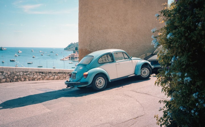 A two-tone blue and white Volkswagen Beetle is parked on a paved road overlooking a bay filled with boats.