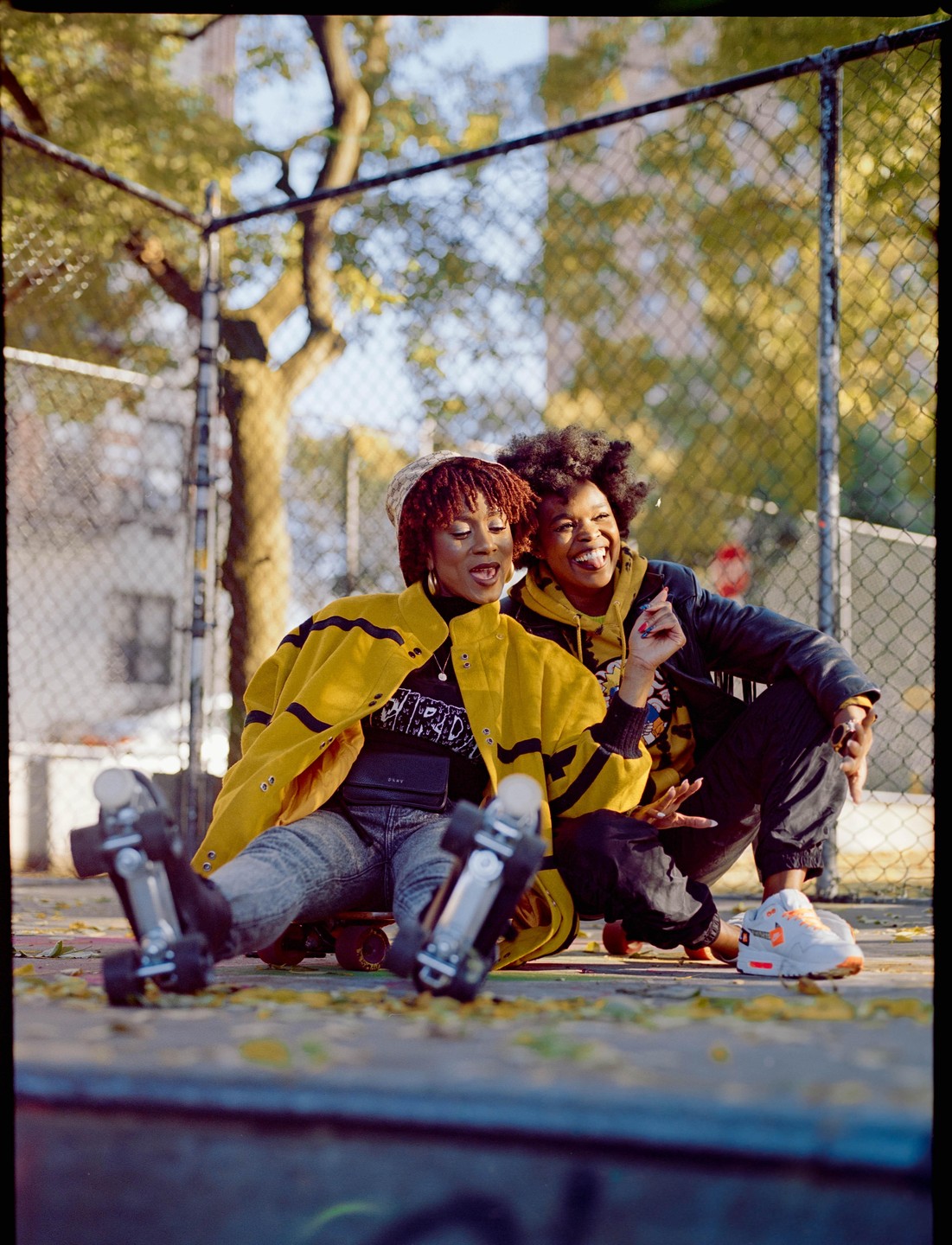 Two women are laughing and smiling while sitting on the ground next to roller skates in a park.