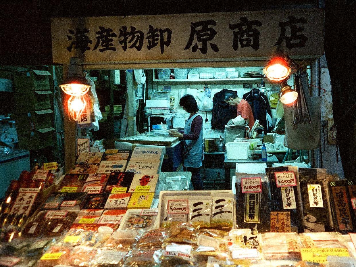 A woman and a man are working inside a small, brightly lit market stall at night, surrounded by various packaged goods.
