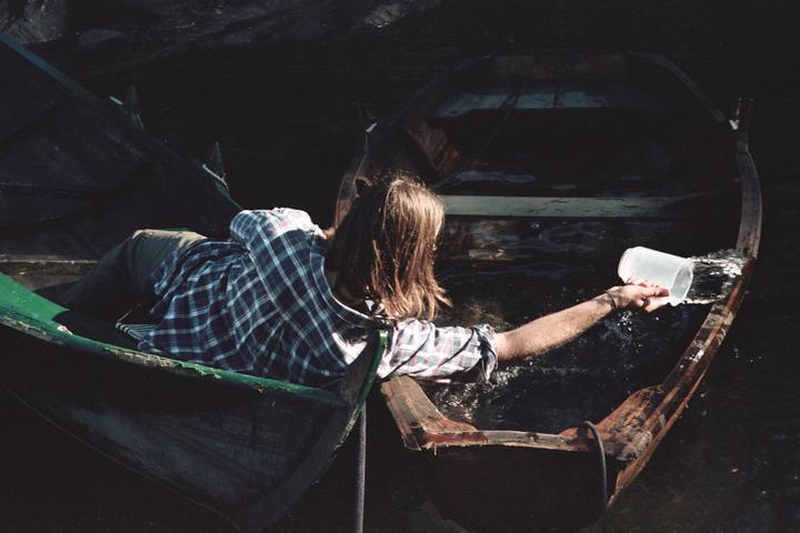 A man in a plaid shirt is bailing water out of a wooden boat with a plastic cup.