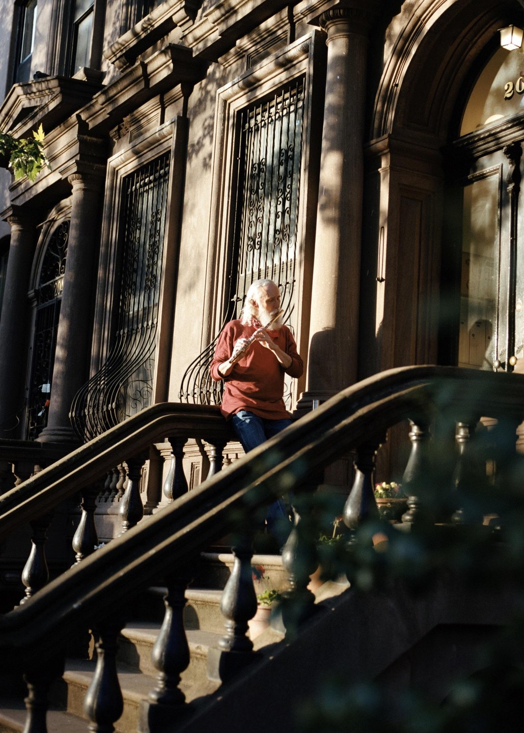 A man with long white hair and beard plays a flute on the steps of a brownstone building in Fort Greene.