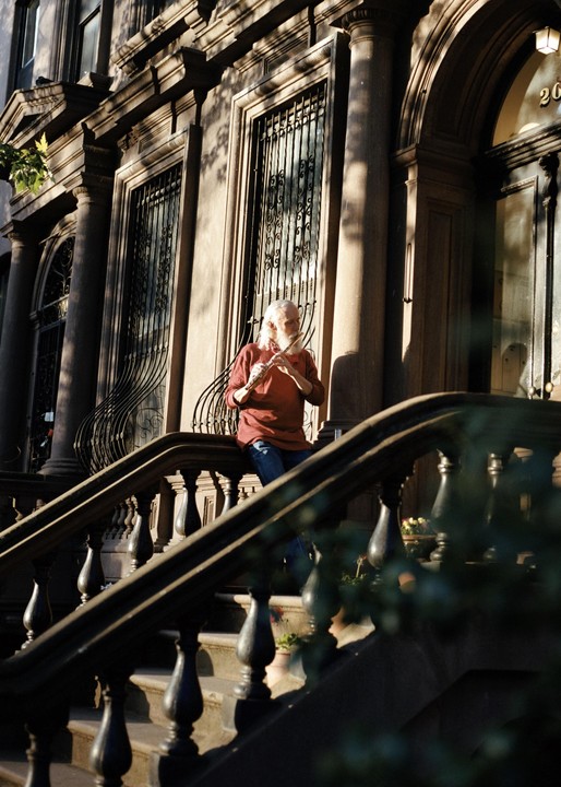 A man with long white hair and beard plays a flute on the steps of a brownstone building in Fort Greene.