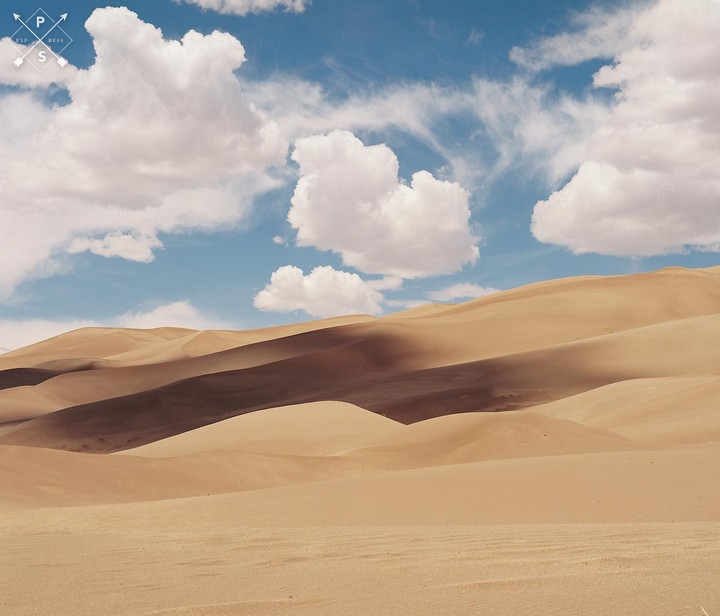 A vast desert landscape features rolling sand dunes under a blue sky with white clouds.