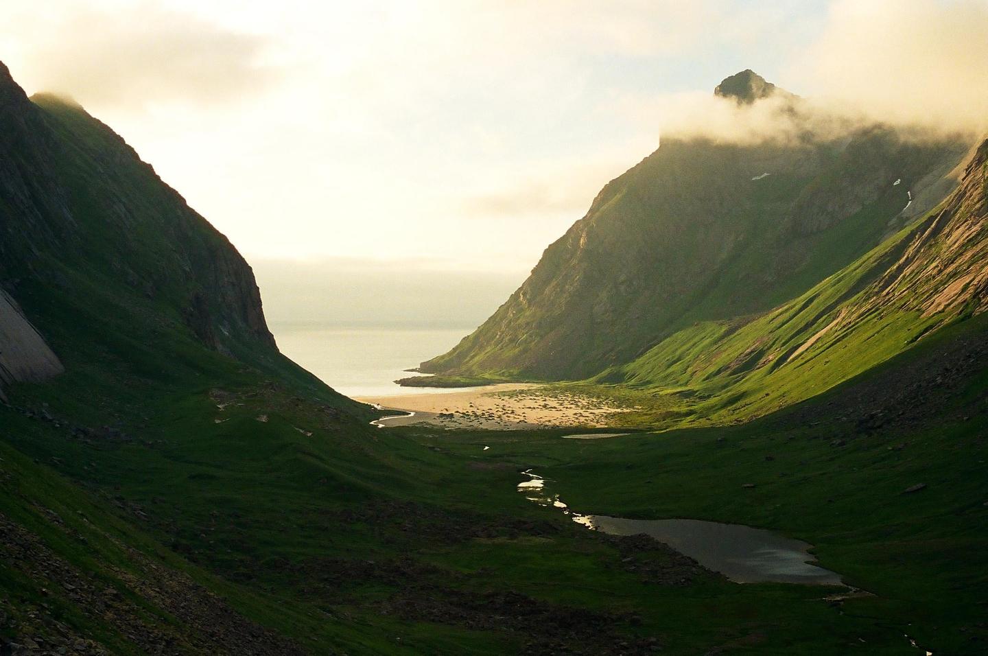 A valley with a stream and a lake leads to a sandy beach and the ocean, flanked by two large mountains under a cloudy sky.