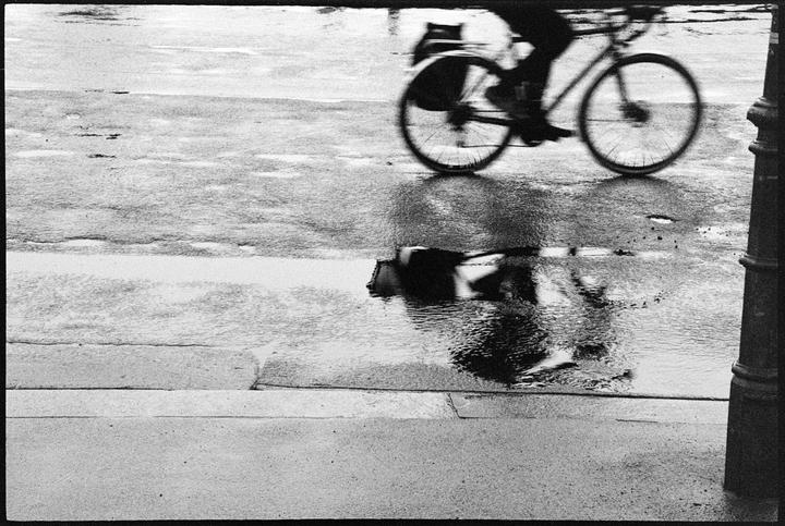 A person rides a bicycle on a wet street, creating a reflection in a puddle.