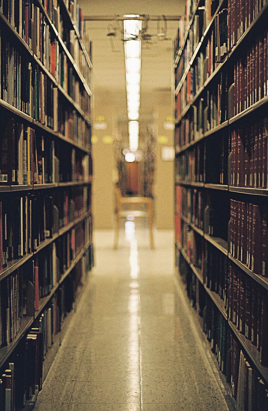 A narrow aisle between tall bookshelves leads to a chair at the end of the library.