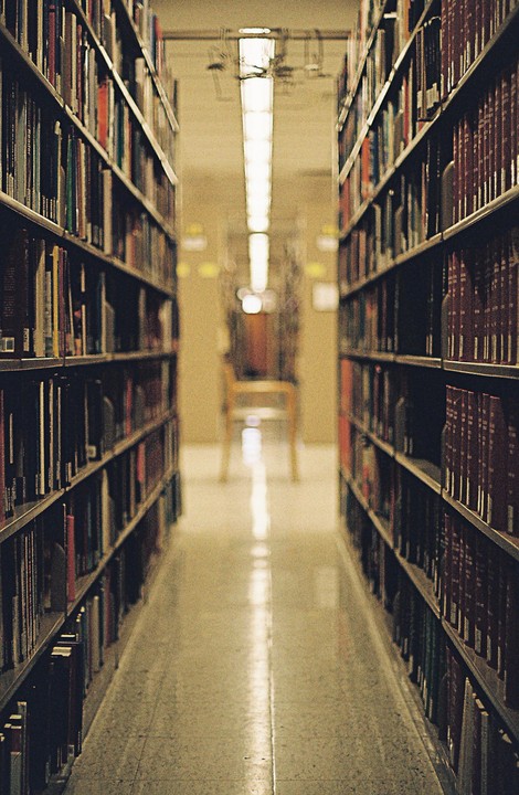 A narrow aisle between tall bookshelves leads to a chair at the end of the library.