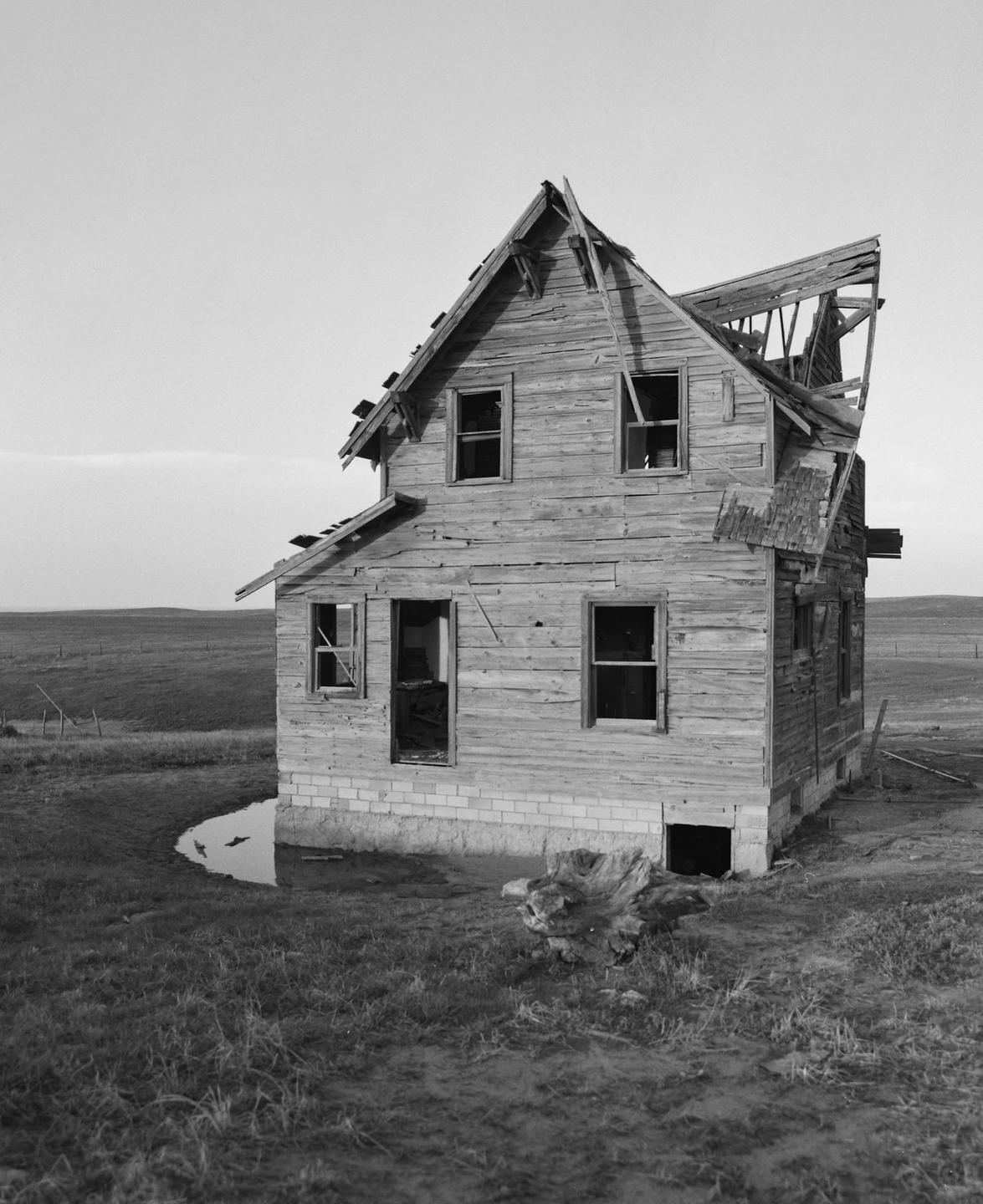 An abandoned wooden house with a partially collapsed roof stands in a grassy field next to a small puddle.