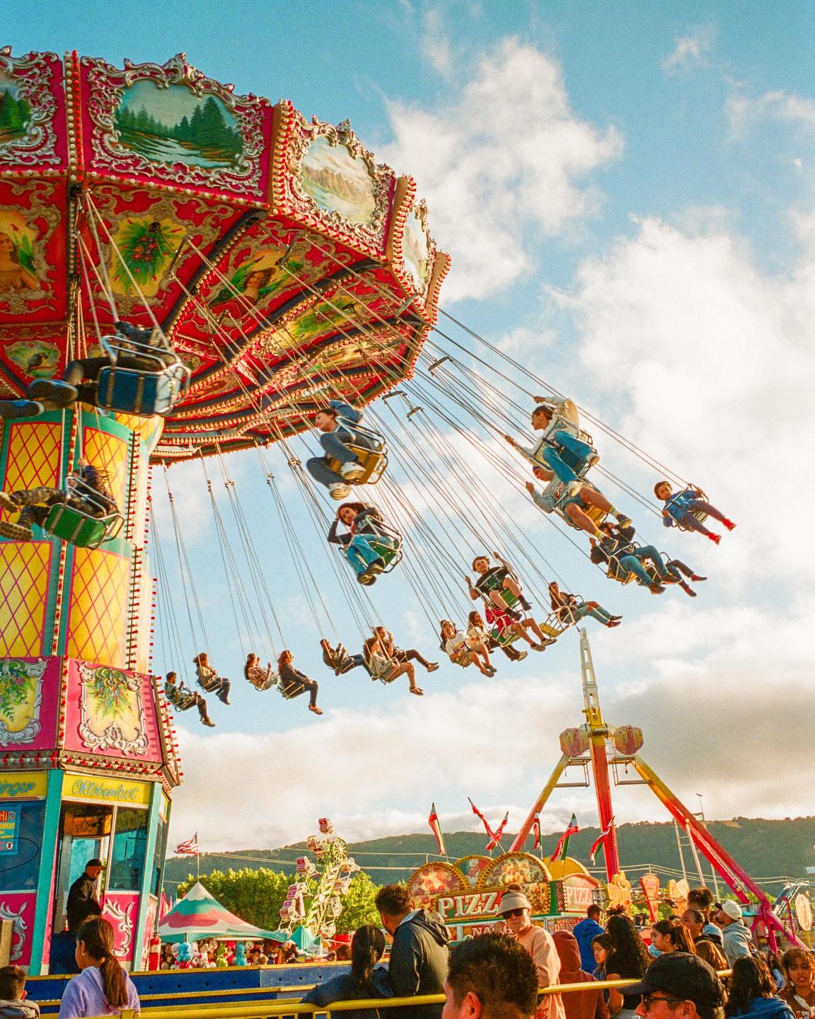 People ride a colorful swing ride at a fairground under a blue sky with clouds.