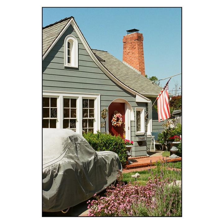 A house with a red door and an American flag has a car covered with a tarp parked in front of it.