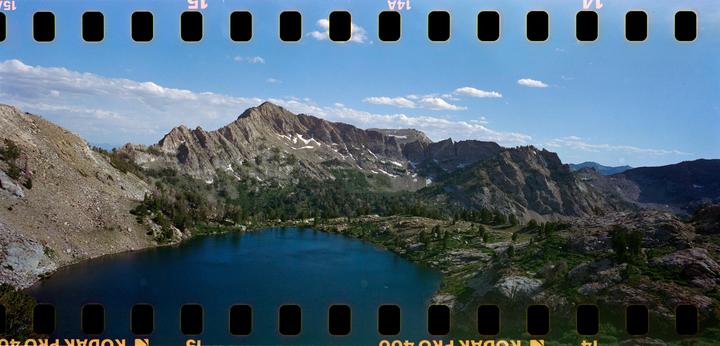 A deep blue alpine lake is surrounded by rocky mountains with patches of snow and green trees under a blue sky with white clouds.
