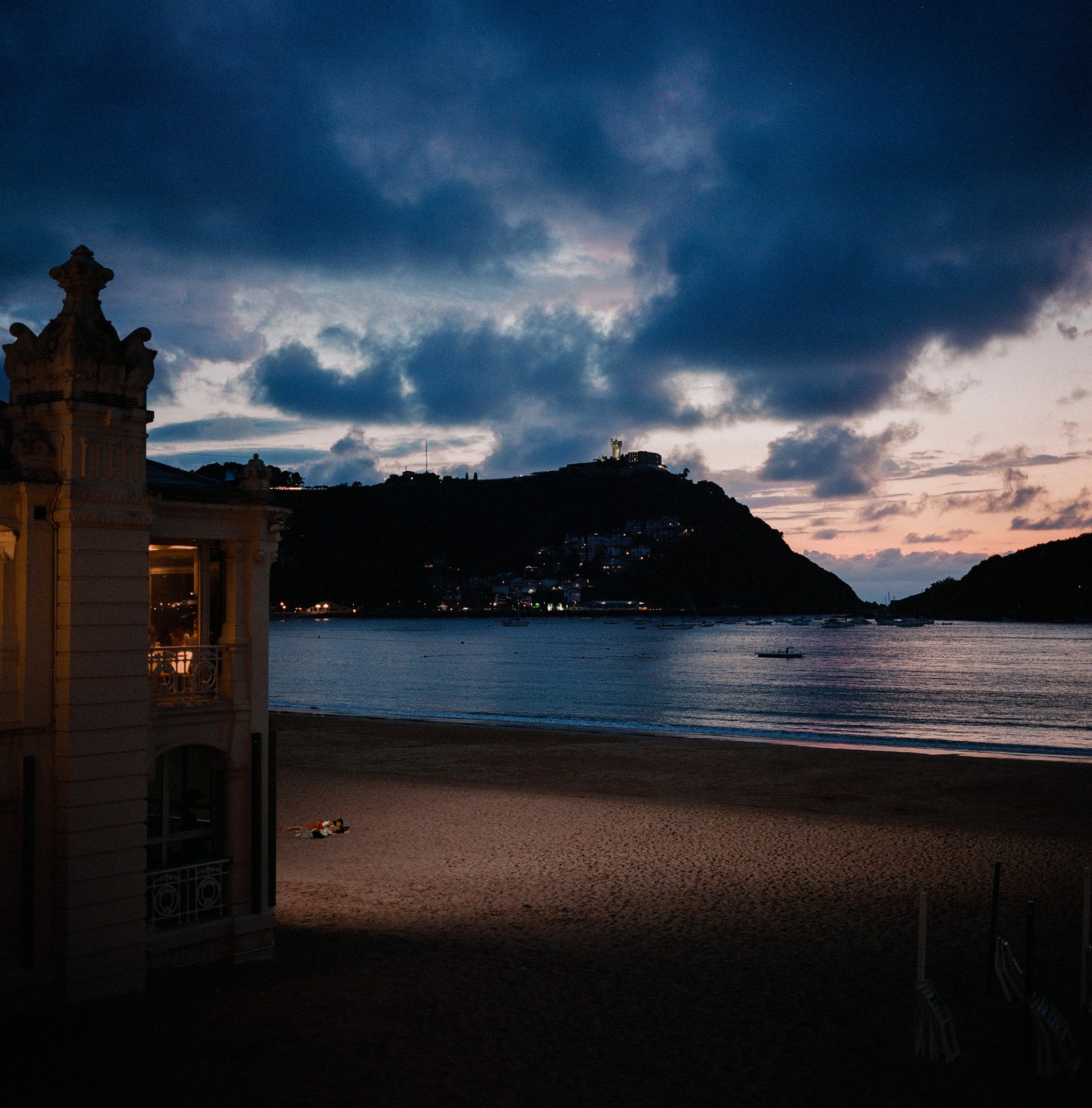 A couple lies on a beach at dusk with a building to the left and a mountain across the water.