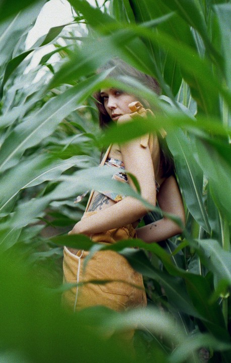 A woman with long hair and a yellow top is partially visible through tall green leaves.