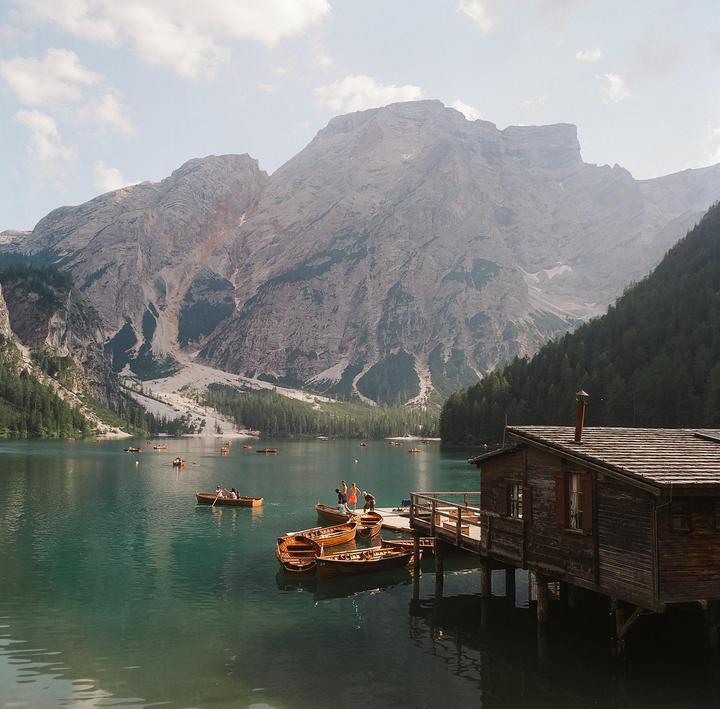 A wooden boathouse and several rowboats are on a lake surrounded by mountains and a forest.