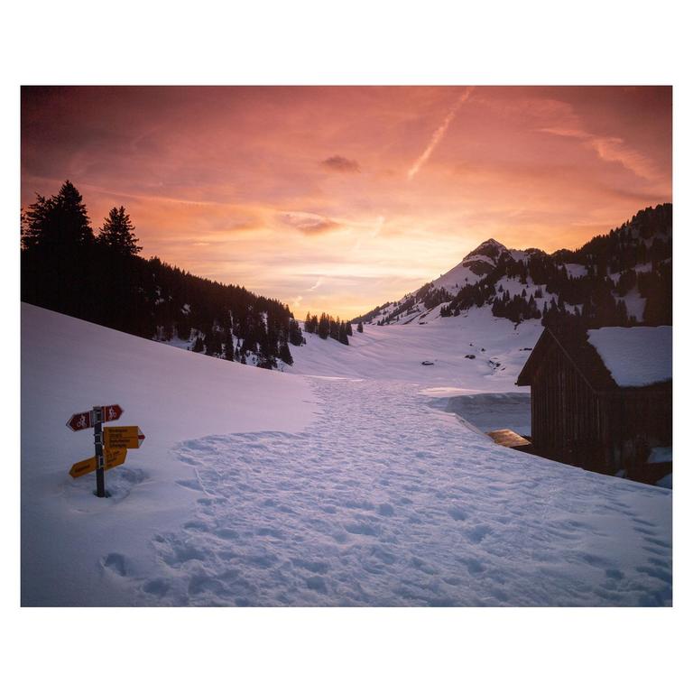 A snow-covered mountain valley with a wooden hut and a signpost is illuminated by the warm glow of sunset.