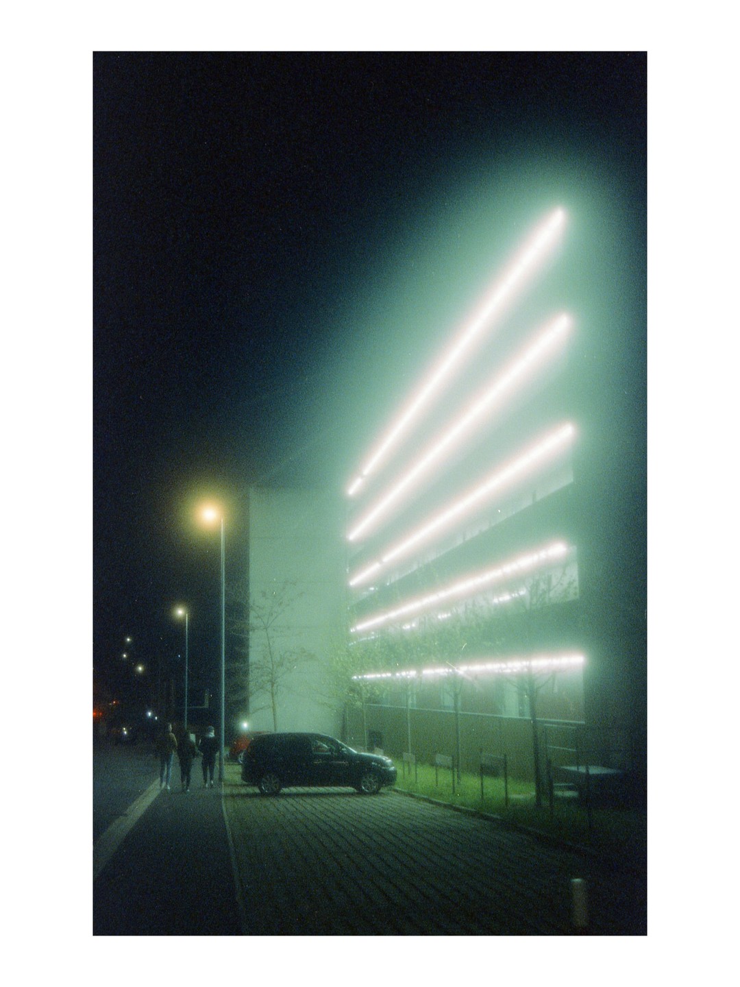 A group of people walk on a sidewalk next to a street light and a parked car, with a building illuminated by bright horizontal lights in the background.