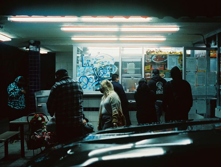 People wait in line at a brightly lit take-out restaurant at night, with a car visible in the foreground.