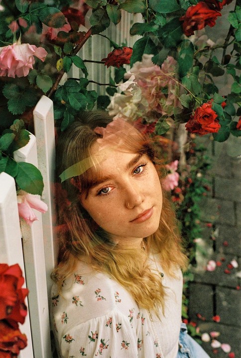 A woman with freckles and a nose ring leans against a white fence, surrounded by red and pink roses.