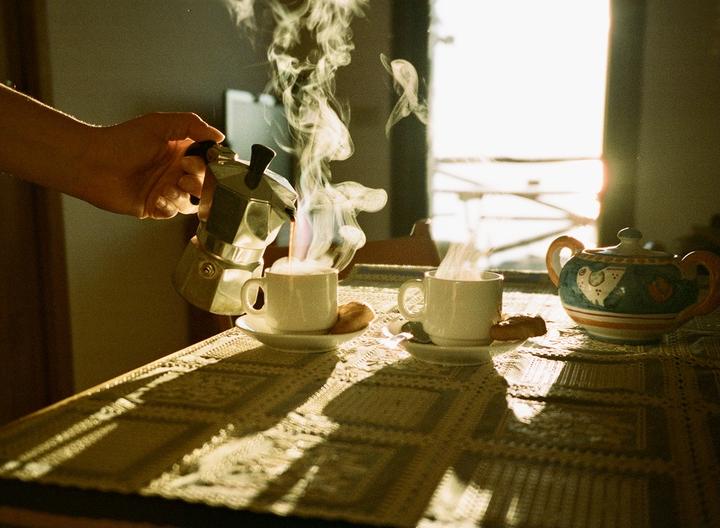 A hand pours coffee from a moka pot into a steaming cup on a table with another cup and a teapot.