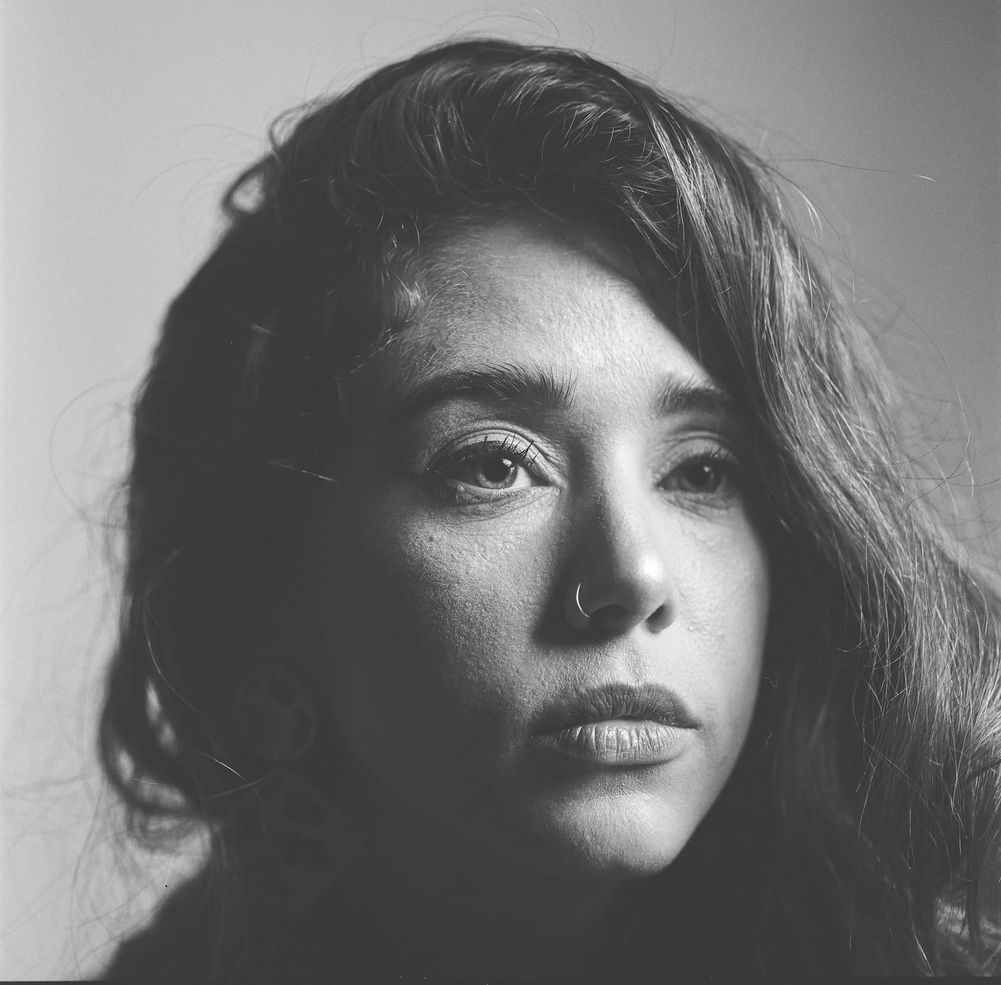 A woman with long hair and a nose ring looks to the side in a close-up portrait.
