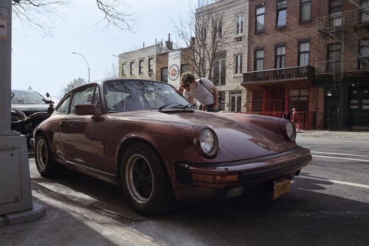 A man wearing sunglasses leans over the hood of a vintage car parked on a city street.