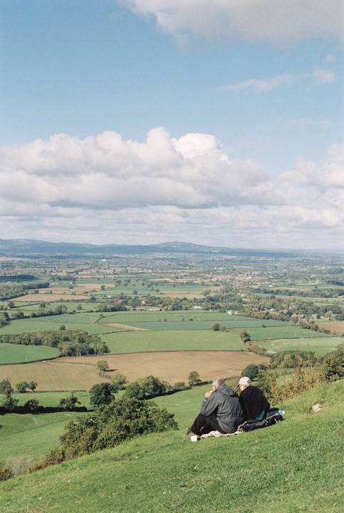 A couple sits on a grassy hill overlooking a vast landscape of fields and distant towns under a cloudy sky.