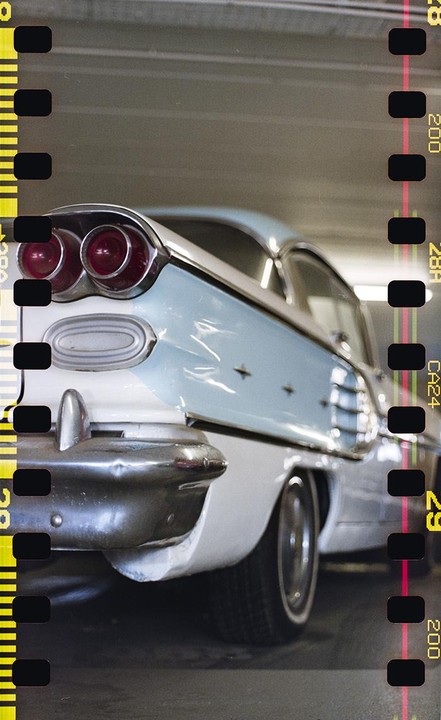 The rear of a light blue and white vintage car with prominent tail lights is visible in a parking garage.