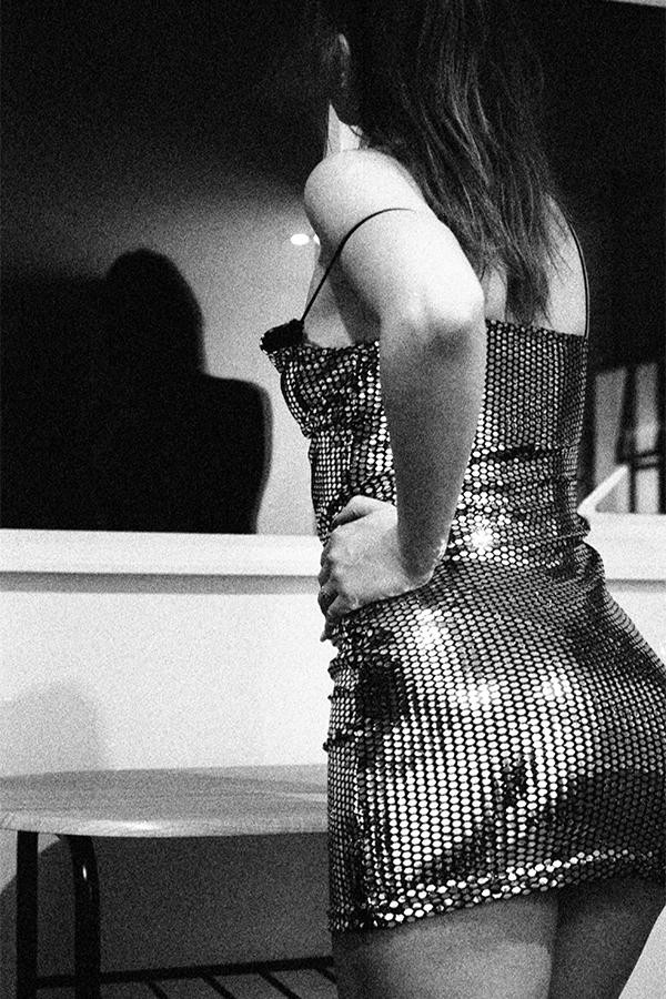 A woman in a sparkly dress stands with her back to the camera, looking at her shadow on a wall.