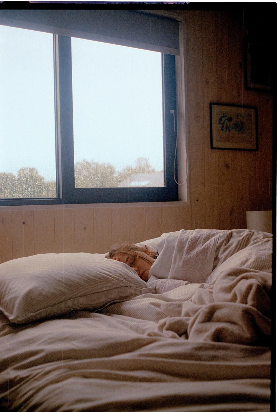 A woman sleeps in a bed next to a window with rain visible outside.