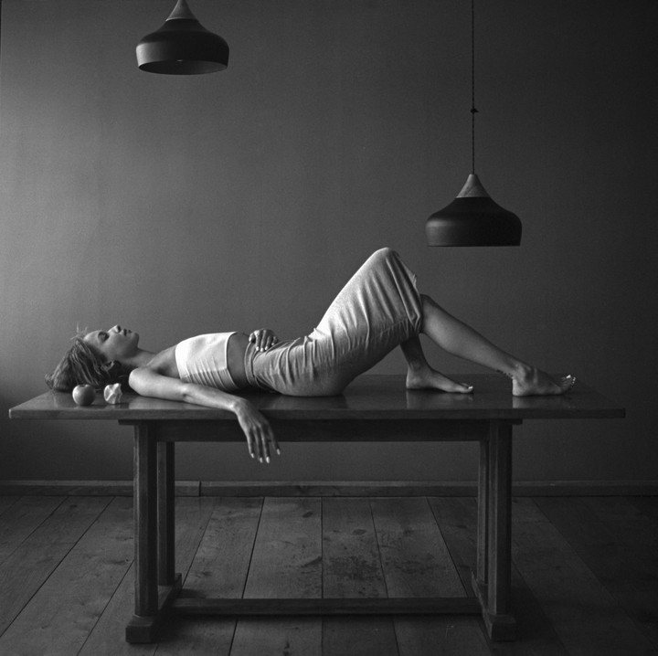 A woman lies on a wooden table with two apples next to her head, under two hanging lamps.