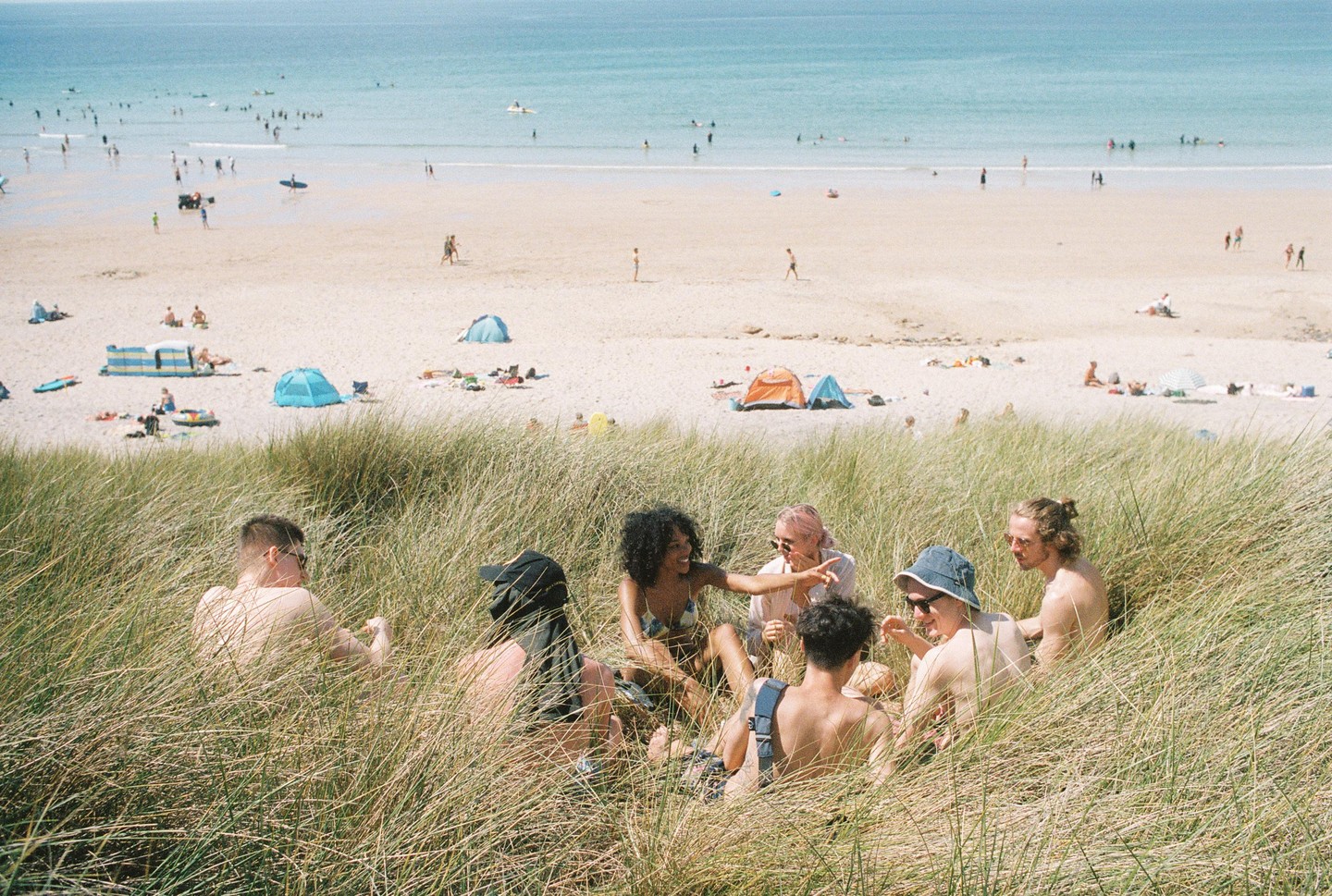 A group of friends sits in tall grass overlooking a crowded beach with people swimming in the ocean.
