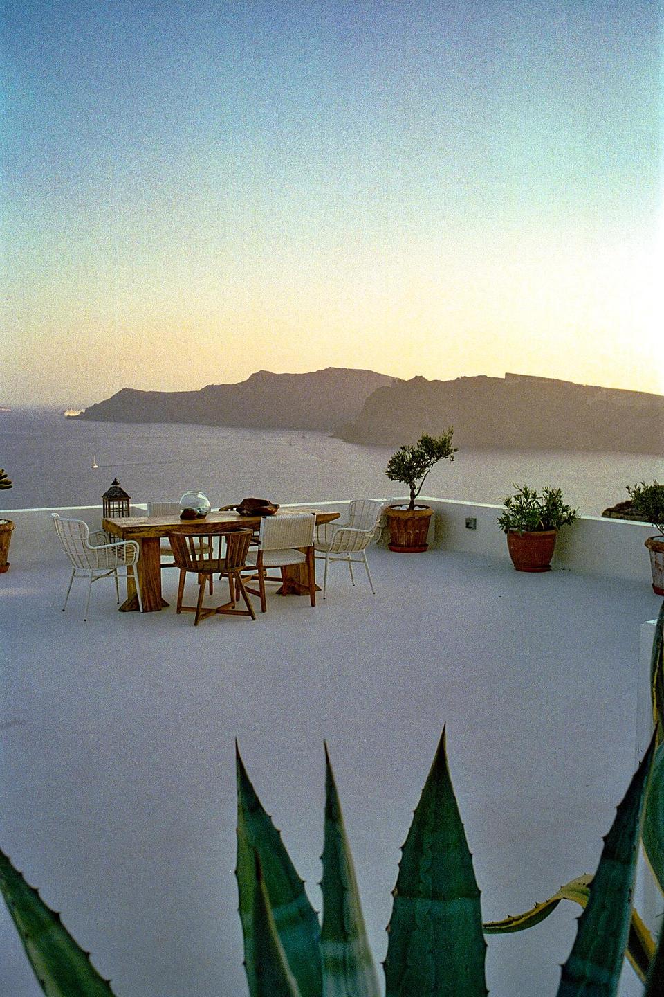 A wooden table and chairs sit on a white patio overlooking a body of water and mountains at sunset.