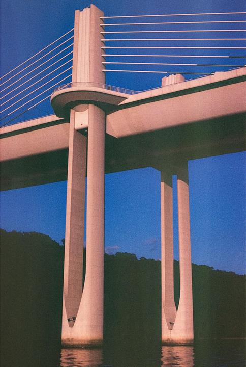 A large concrete bridge with cables is seen from below, with a dark hillside and water visible beneath it.