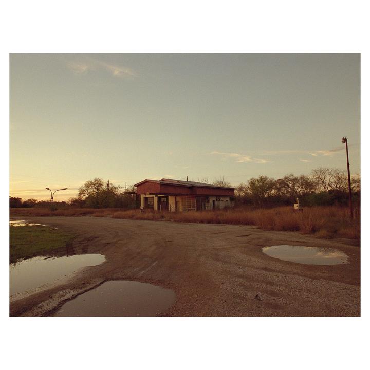 An abandoned building with a red roof stands in a field with puddles at dusk.