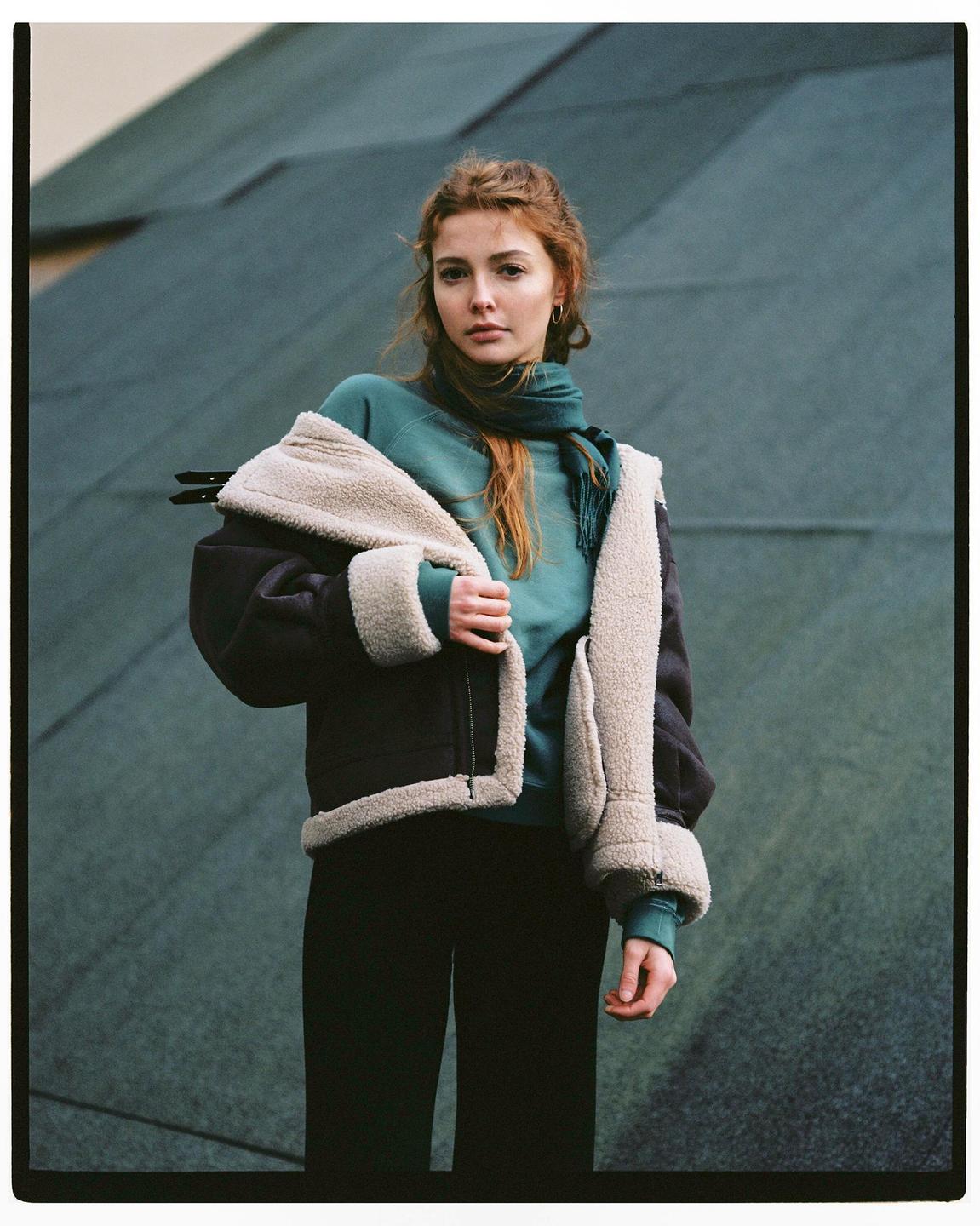 A woman with red hair wears a teal sweatshirt and a black and cream jacket.