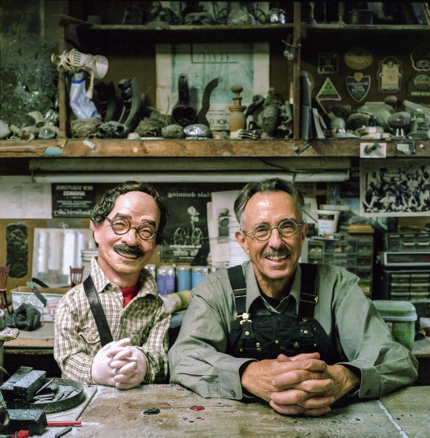 A man and a puppet sit at a messy workbench in a workshop.