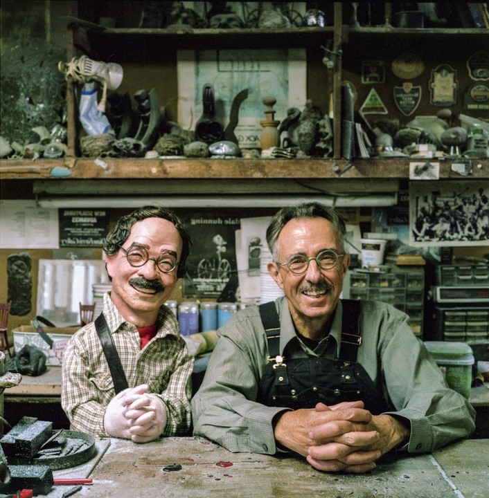 A man and a puppet sit at a messy workbench in a workshop.