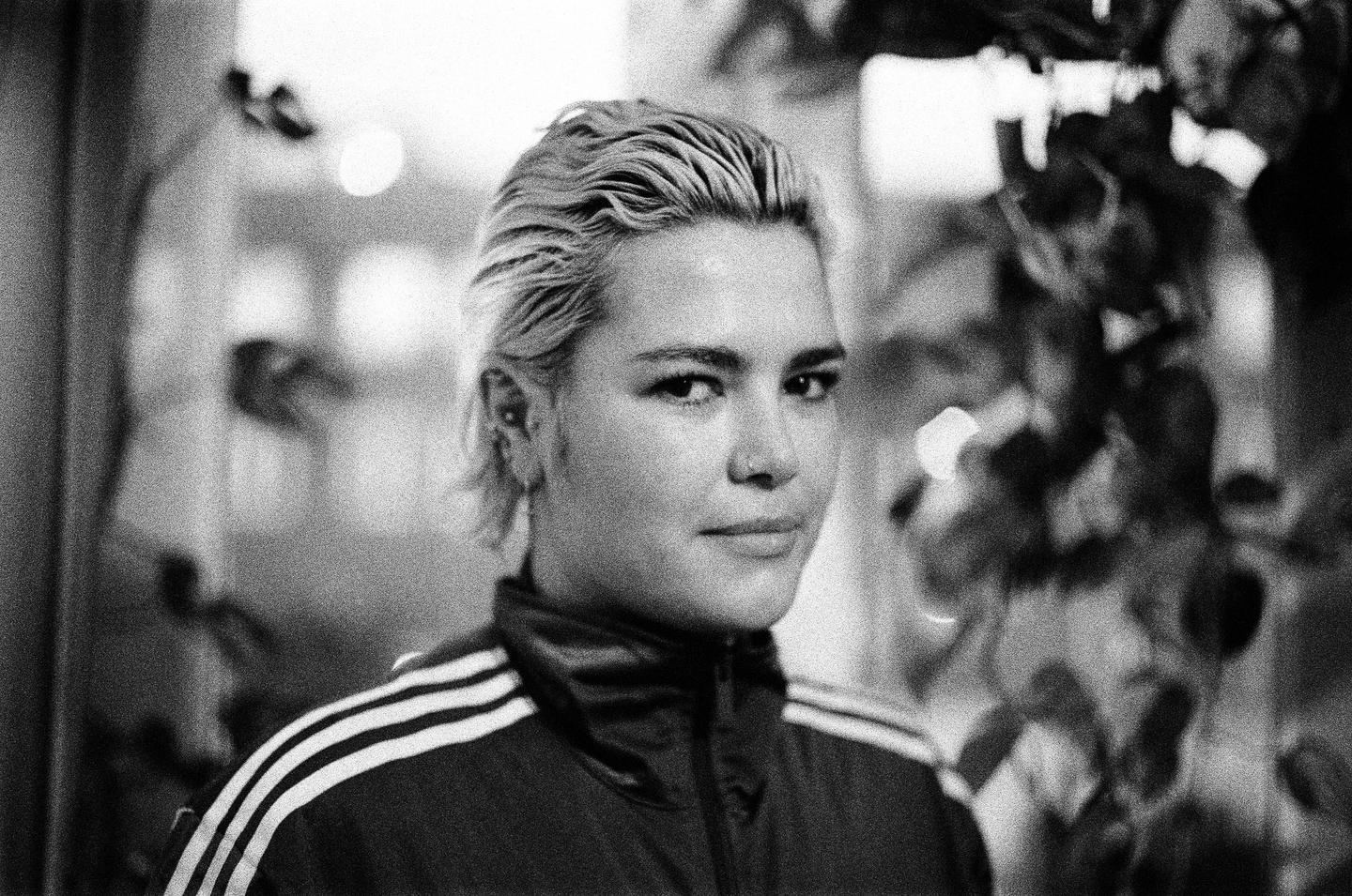 A woman with short, light-colored hair and a track jacket looks towards the viewer.