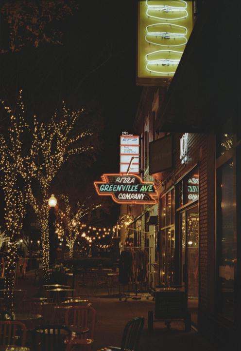 A street at night is illuminated by neon signs and string lights wrapped around trees.