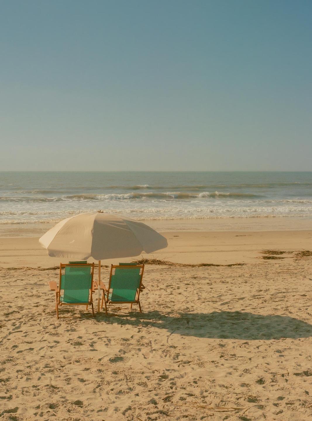 Two beach chairs sit under an umbrella on a sandy beach with the ocean in the background.