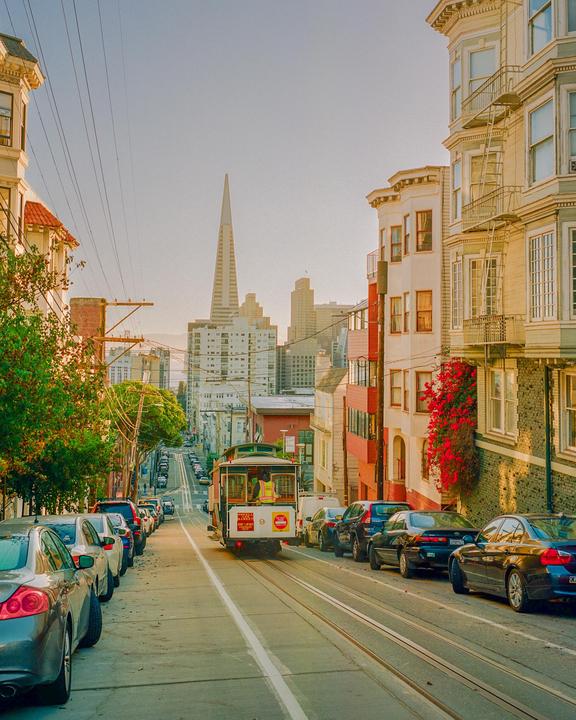 A cable car travels down a steep street lined with parked cars and buildings, with a tall, pointed skyscraper in the distance.