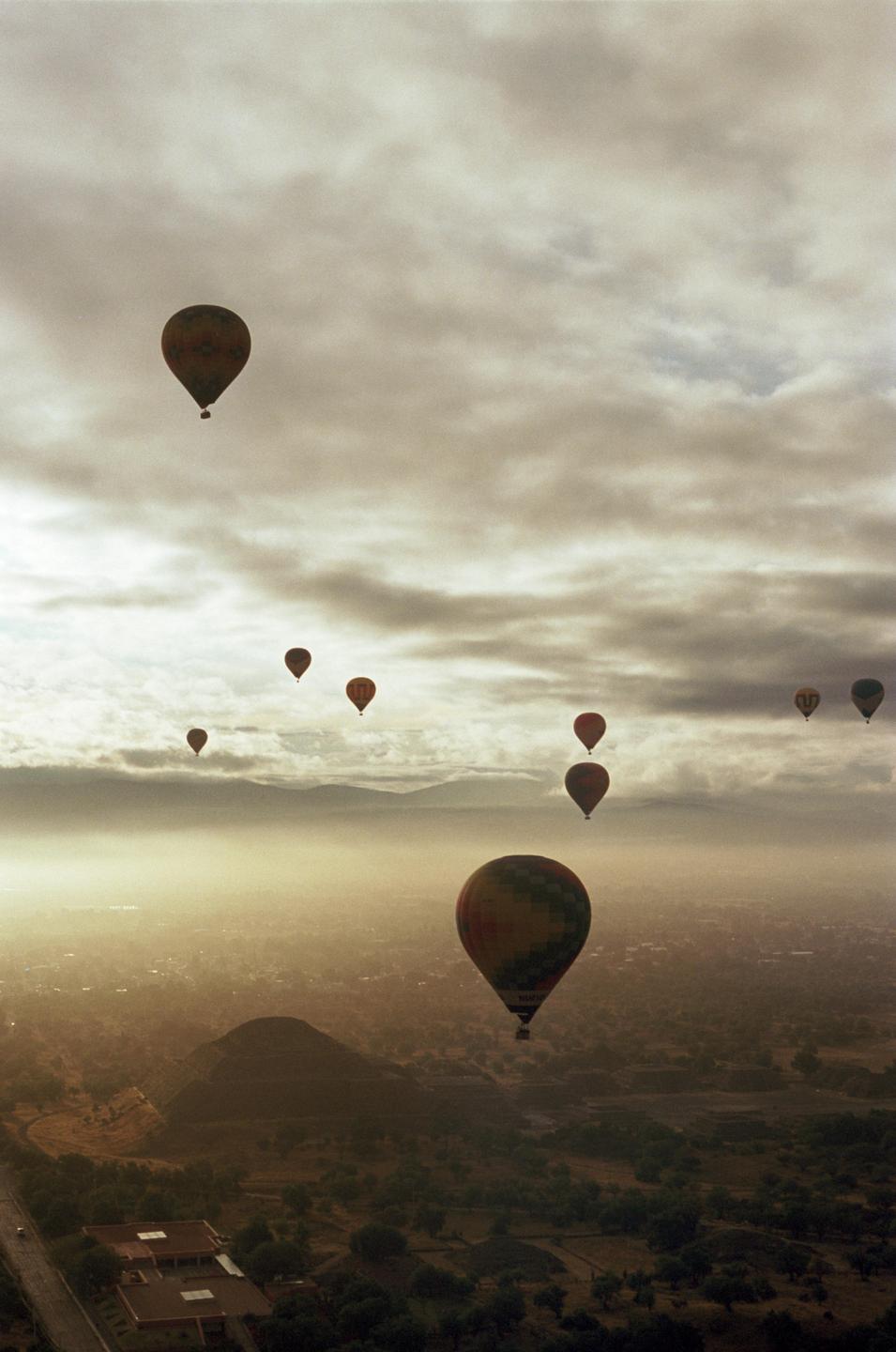 Hot air balloons float above a hazy landscape with a pyramid and mountains under a cloudy sky.
