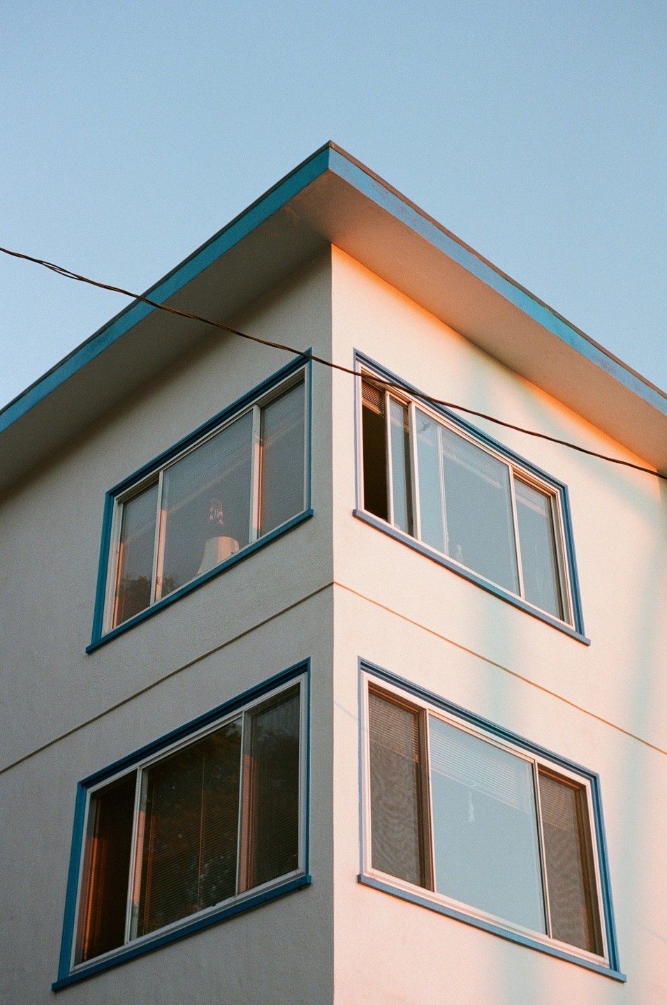 The corner of a building with blue trim and multiple windows is illuminated by the golden hour light.