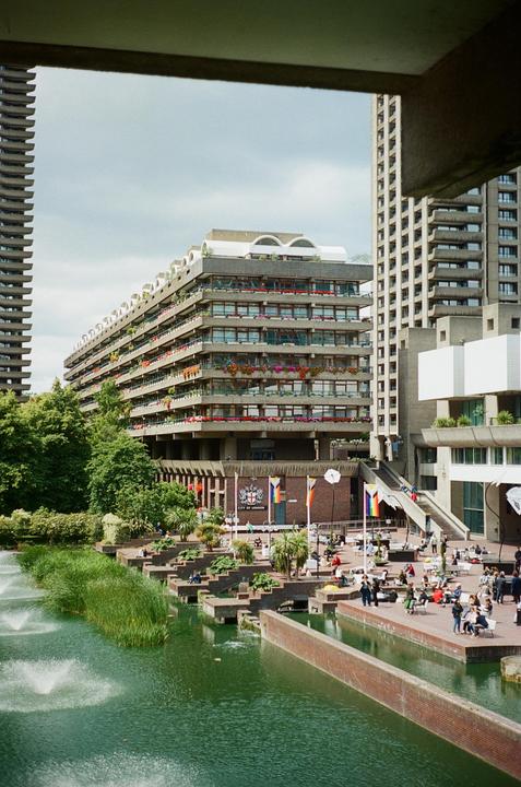 A brutalist building with balconies adorned with flowers overlooks a pond with fountains and people relaxing on a sunny day.