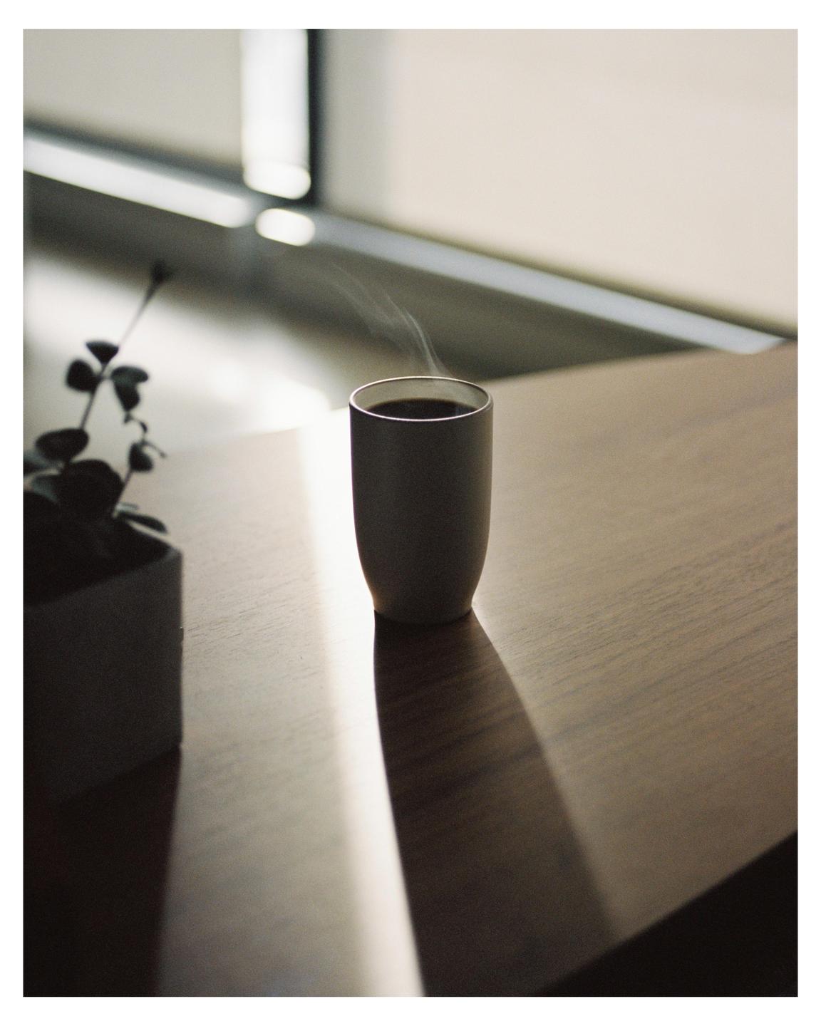 A steaming cup of coffee sits on a wooden table next to a potted plant, with a window in the background.