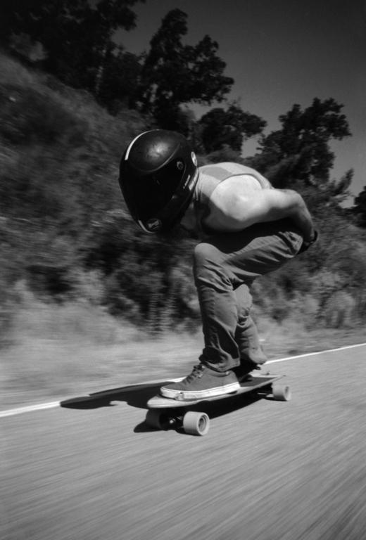 A man wearing a helmet and jeans is skateboarding down a road with trees in the background.