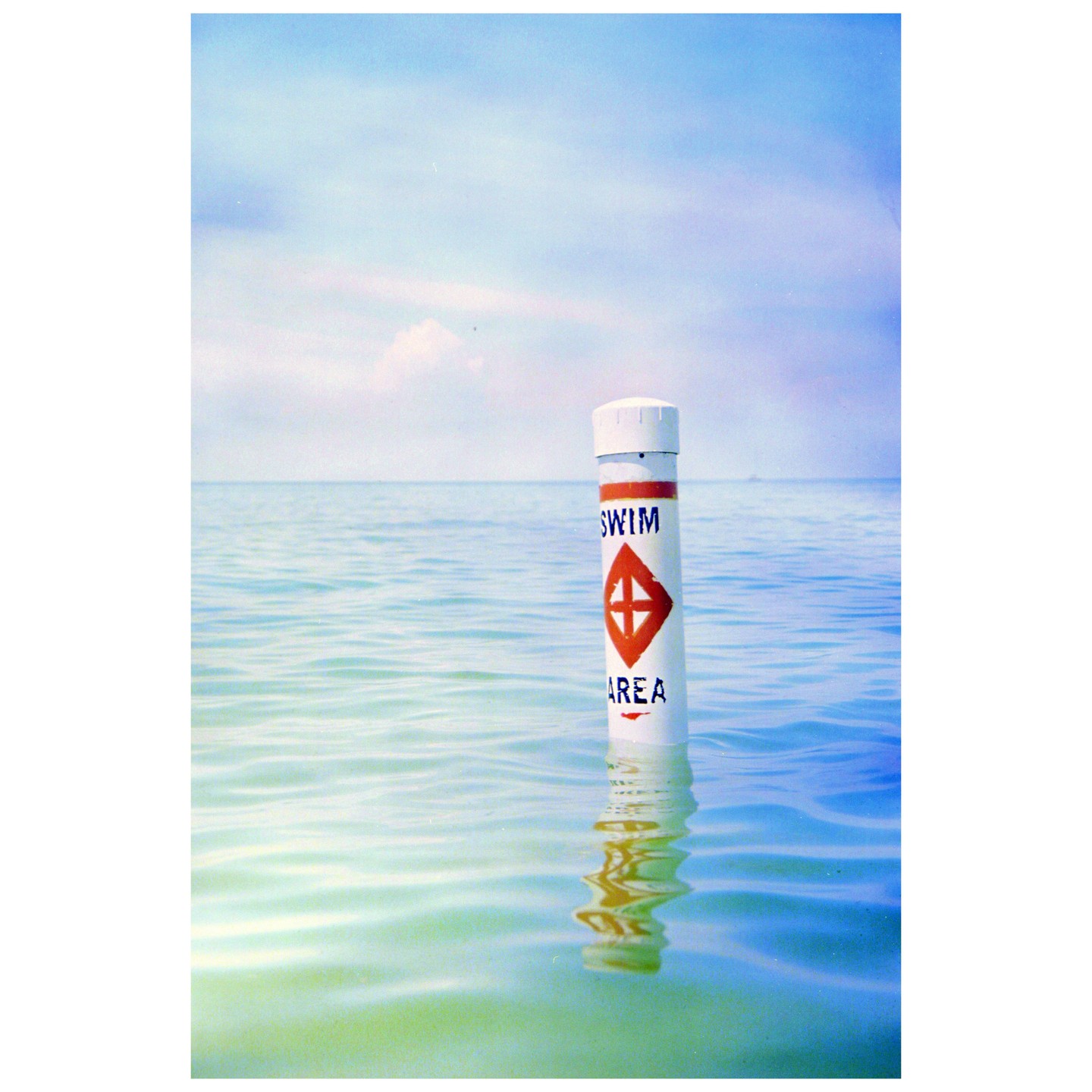 A white buoy with "SWIM AREA" written on it floats in calm water under a light blue sky.