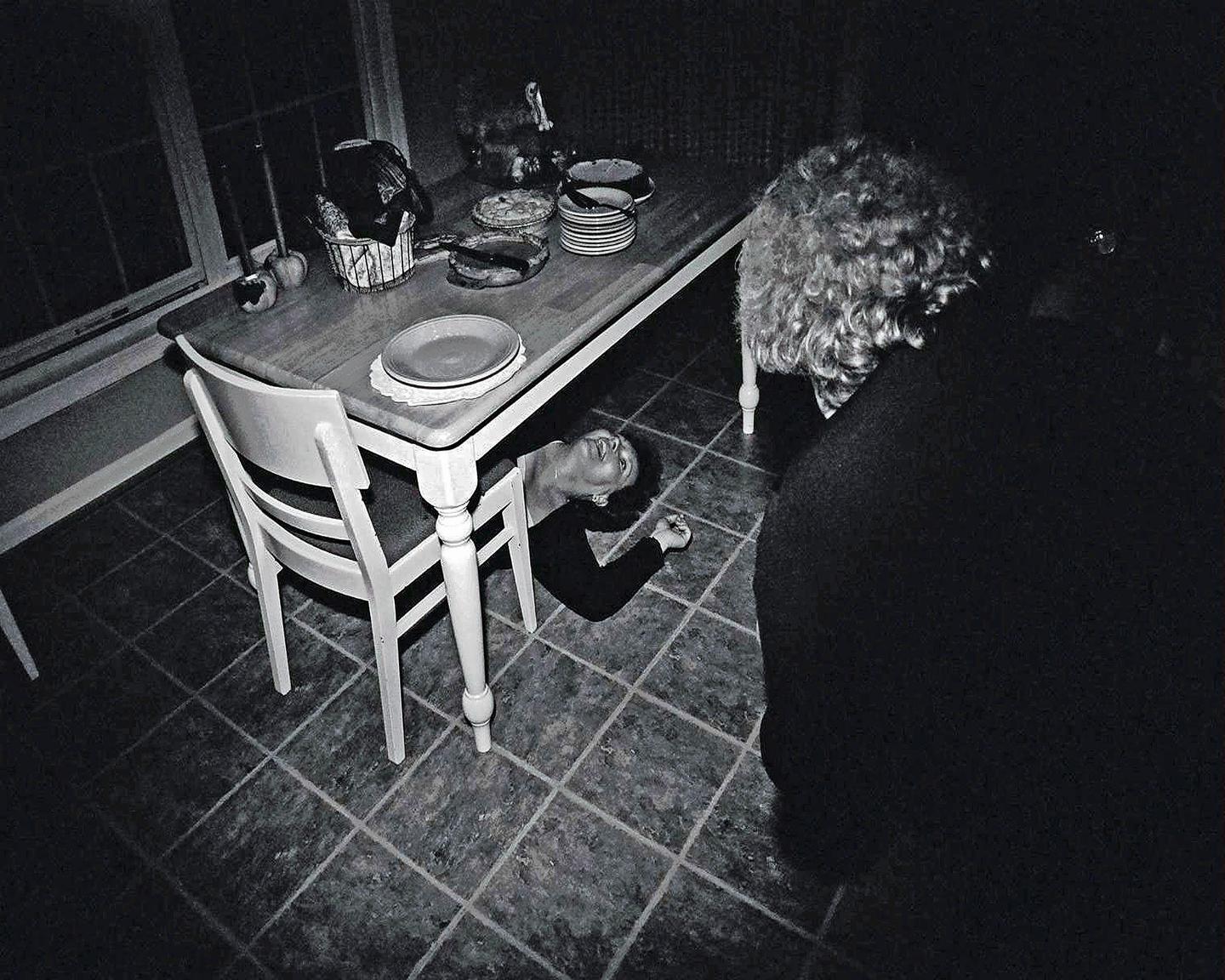 A woman lies on the floor under a dining table while another person stands nearby.