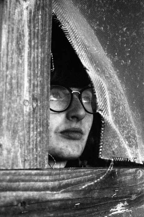 A woman wearing glasses peeks through a torn screen next to a wooden post.