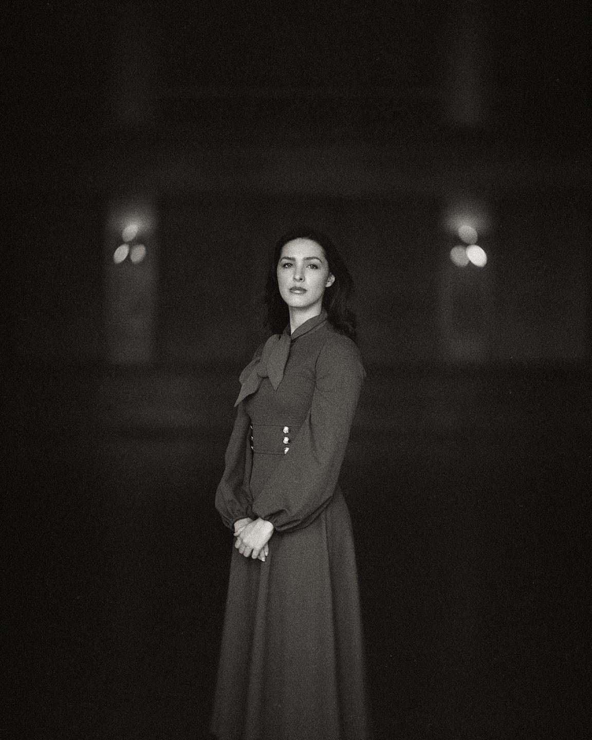 A woman in a long dress stands with her hands clasped in front of her, looking directly at the viewer.